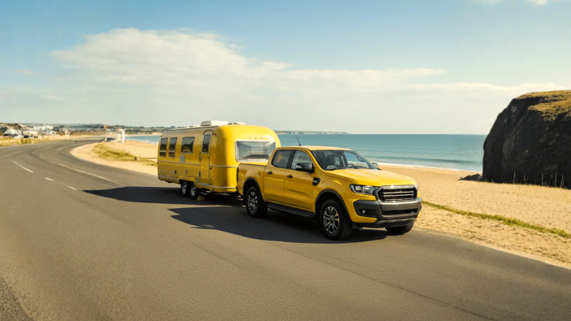 A yellow ute towing a caravan along a coastal road.